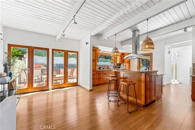 kitchen featuring beamed ceiling, pendant lighting, light wood-style floors, island range hood, and brown cabinetry