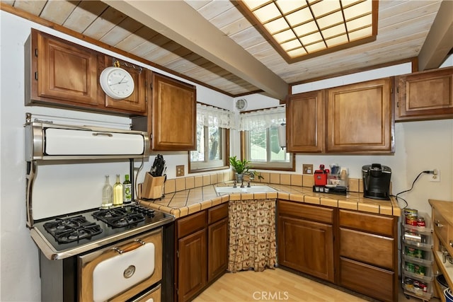 kitchen featuring tile countertops, brown cabinetry, stainless steel gas range, beam ceiling, and a sink