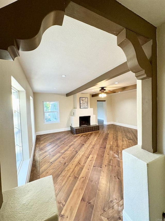 unfurnished living room with beam ceiling, a tiled fireplace, wood-type flooring, baseboards, and ceiling fan