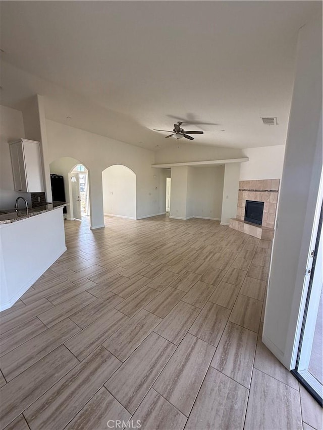 unfurnished living room featuring a tile fireplace, ceiling fan, visible vents, wood finish floors, and arched walkways