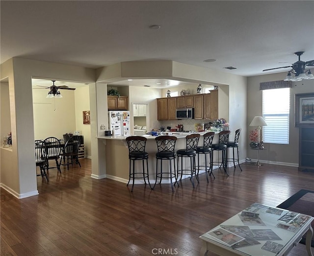 kitchen featuring stainless steel microwave, dark wood-type flooring, freestanding refrigerator, brown cabinetry, and a ceiling fan