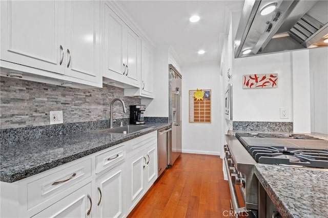 kitchen featuring wall chimney range hood, decorative backsplash, appliances with stainless steel finishes, white cabinets, and a sink