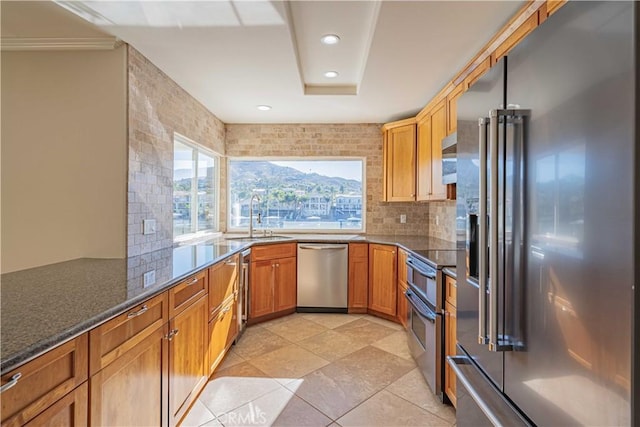 kitchen with brown cabinetry, dark stone counters, a sink, decorative backsplash, and stainless steel appliances