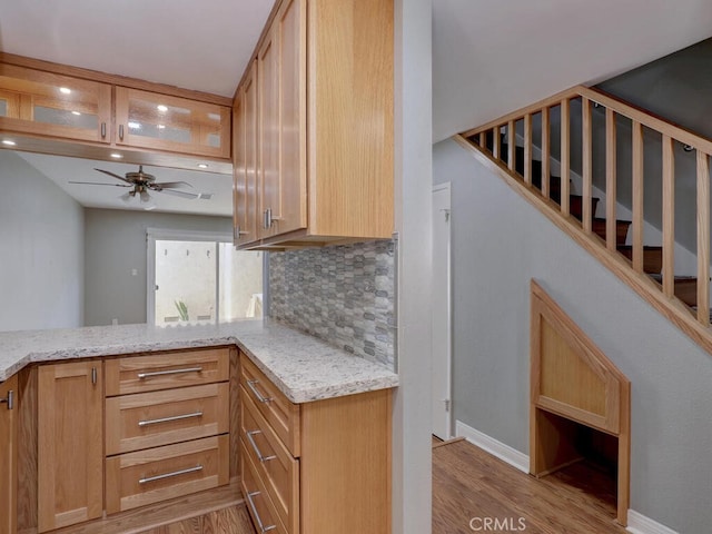 kitchen with light wood finished floors, glass insert cabinets, ceiling fan, light stone countertops, and decorative backsplash