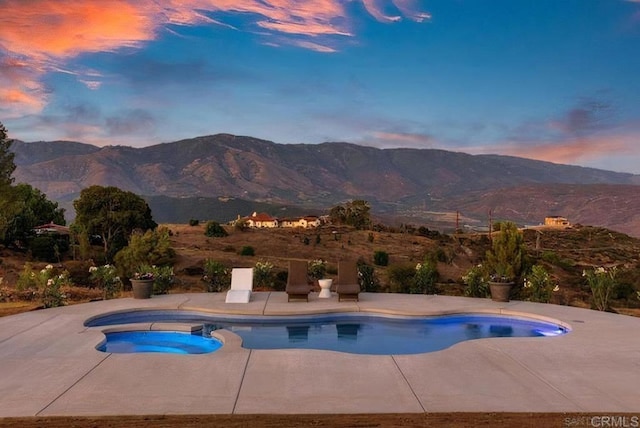 pool at dusk featuring a mountain view, a pool with connected hot tub, and a patio area