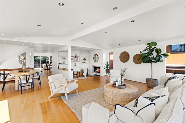 living area with built in shelves, lofted ceiling with beams, a lit fireplace, recessed lighting, and light wood-style flooring