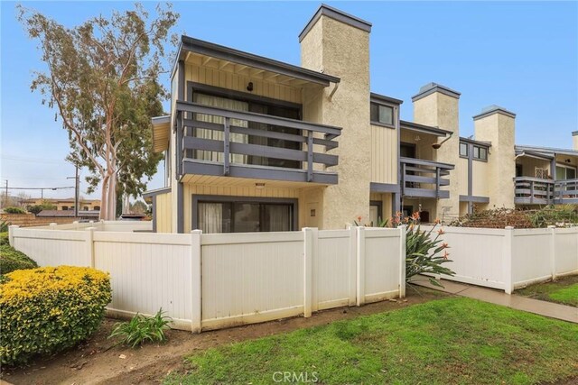 rear view of house featuring stucco siding, a balcony, a chimney, and fence