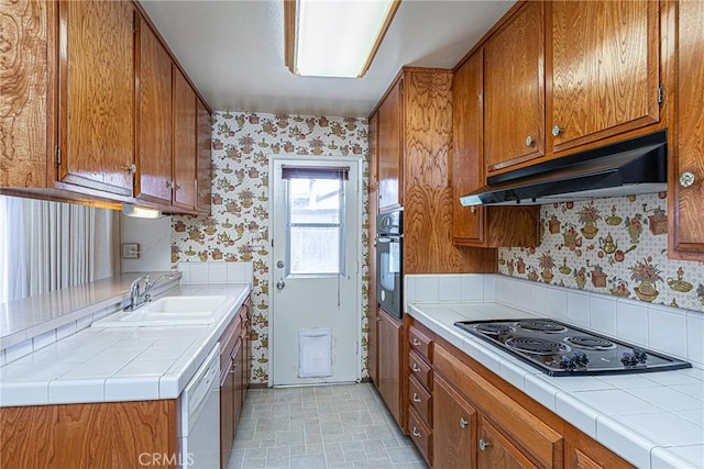 kitchen with black appliances, under cabinet range hood, a sink, brown cabinetry, and wallpapered walls