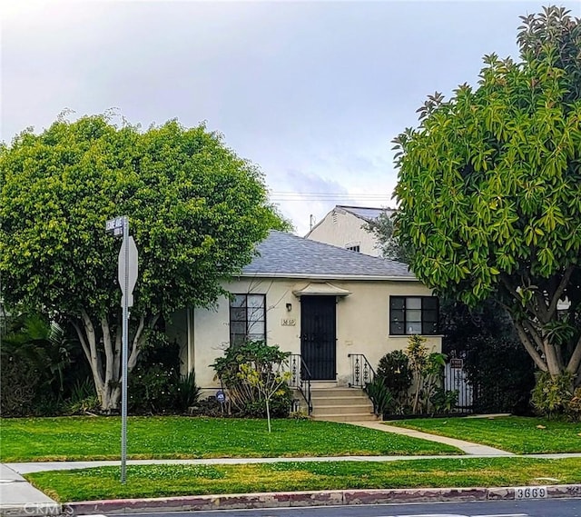 view of front facade with stucco siding, roof with shingles, a front yard, and fence