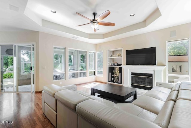 living area with ceiling fan, a raised ceiling, built in shelves, and wood finished floors