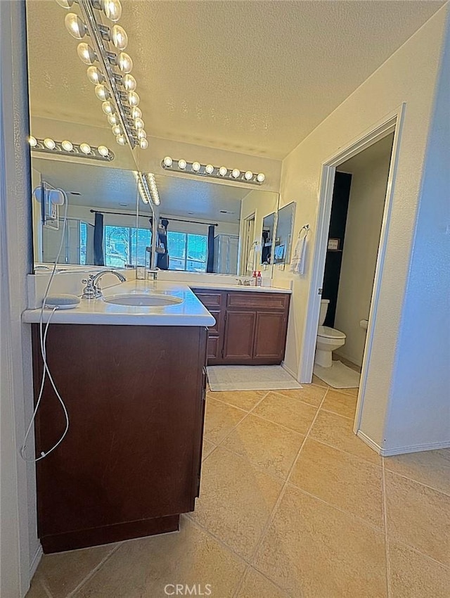 bathroom with baseboards, toilet, two vanities, a textured ceiling, and a sink