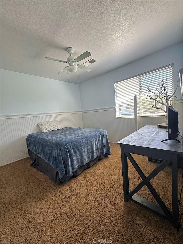 carpeted bedroom featuring a textured ceiling, a ceiling fan, visible vents, and wainscoting