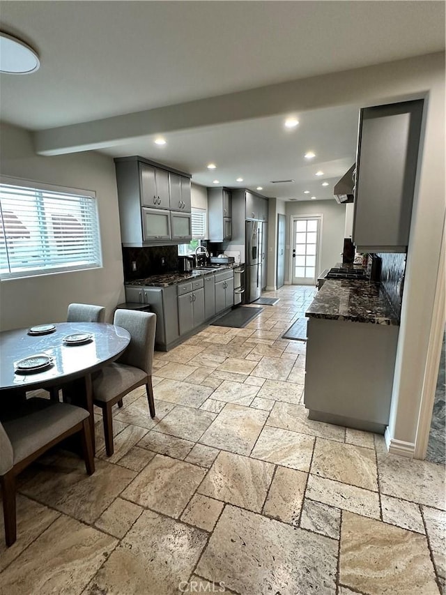 kitchen with gray cabinetry, stainless steel fridge with ice dispenser, stone tile floors, recessed lighting, and wall chimney exhaust hood