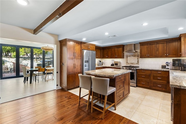 kitchen featuring high end appliances, a kitchen island, a tray ceiling, wall chimney range hood, and tasteful backsplash