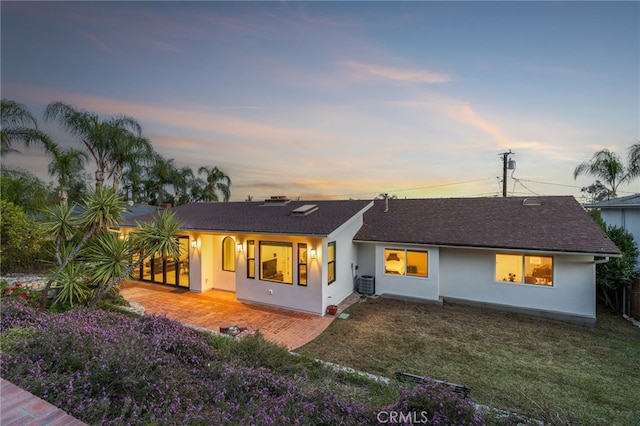 back of house at dusk with central air condition unit, stucco siding, a lawn, a patio, and roof with shingles