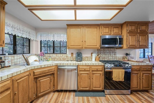 kitchen with tile countertops, brown cabinetry, stainless steel appliances, light wood-style floors, and backsplash