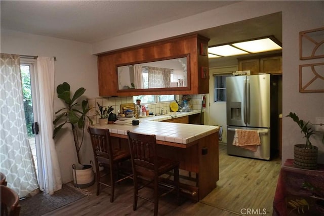 kitchen featuring light wood finished floors, tasteful backsplash, stainless steel fridge with ice dispenser, tile countertops, and a peninsula