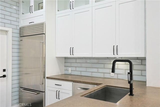 kitchen with a sink, stainless steel built in fridge, white cabinets, glass insert cabinets, and backsplash