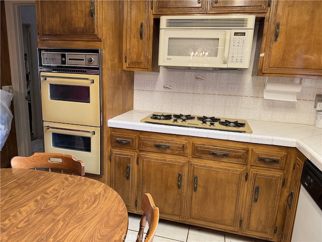 kitchen with tile countertops, decorative backsplash, brown cabinets, light tile patterned flooring, and white appliances