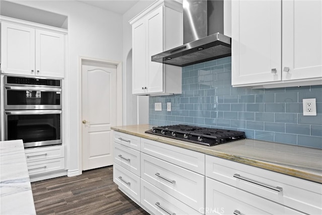 kitchen featuring dark wood-style floors, stainless steel double oven, white cabinets, gas stovetop, and wall chimney exhaust hood