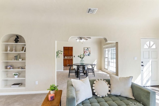 carpeted living room featuring visible vents, built in shelves, ceiling fan, baseboards, and arched walkways