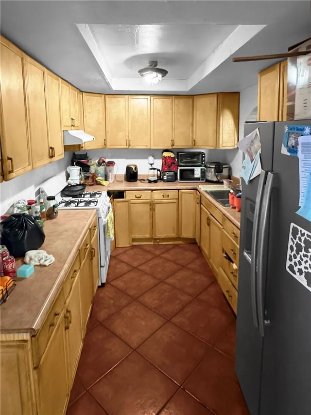 kitchen with under cabinet range hood, dark tile patterned floors, white range with gas cooktop, stainless steel fridge, and a raised ceiling