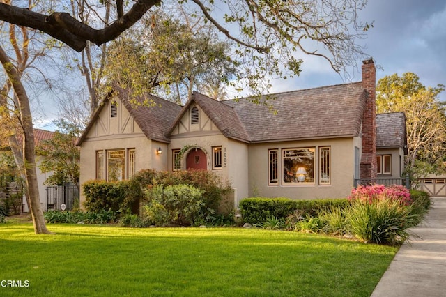 tudor home with a front lawn, fence, roof with shingles, stucco siding, and a chimney