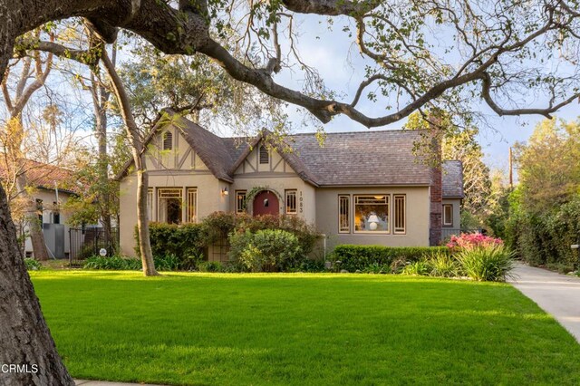 english style home with a shingled roof, a front yard, fence, and stucco siding