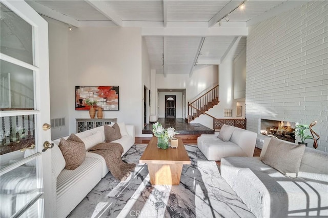 living area featuring track lighting, beamed ceiling, stairs, a fireplace, and a towering ceiling