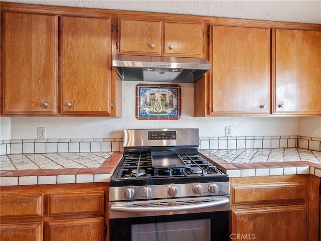 kitchen featuring under cabinet range hood, brown cabinetry, tile countertops, and stainless steel range with gas cooktop