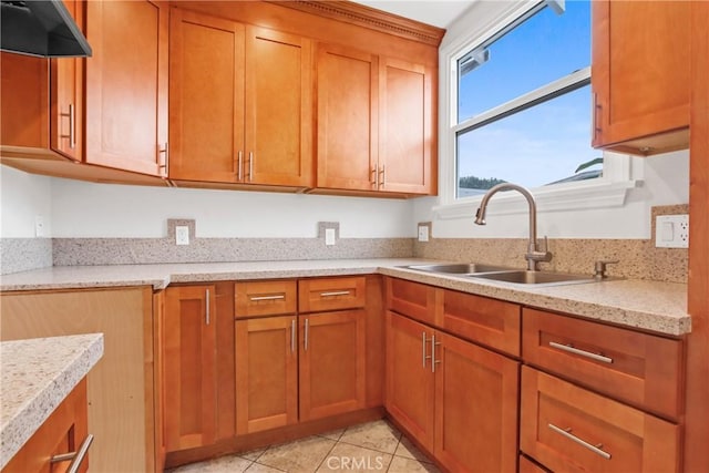 kitchen with a sink, under cabinet range hood, brown cabinetry, light tile patterned floors, and light stone countertops