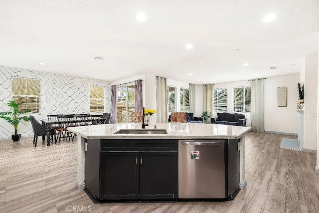 kitchen featuring stainless steel dishwasher, open floor plan, a kitchen island with sink, light wood-style flooring, and dark cabinetry