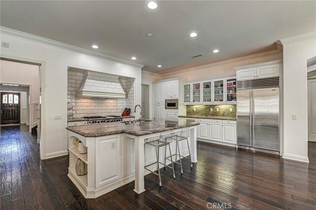 kitchen featuring built in refrigerator, crown molding, a center island with sink, custom exhaust hood, and a breakfast bar area