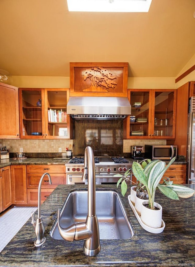 kitchen with backsplash, appliances with stainless steel finishes, glass insert cabinets, and wall chimney range hood