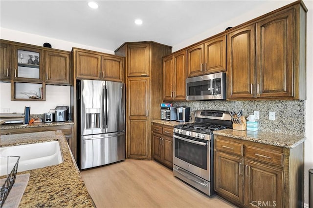 kitchen featuring tasteful backsplash, stainless steel appliances, light wood-style floors, glass insert cabinets, and light stone countertops