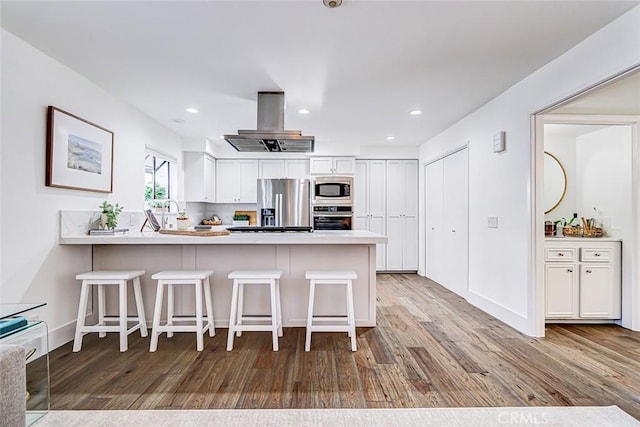 kitchen with wood finished floors, appliances with stainless steel finishes, a peninsula, island range hood, and white cabinets