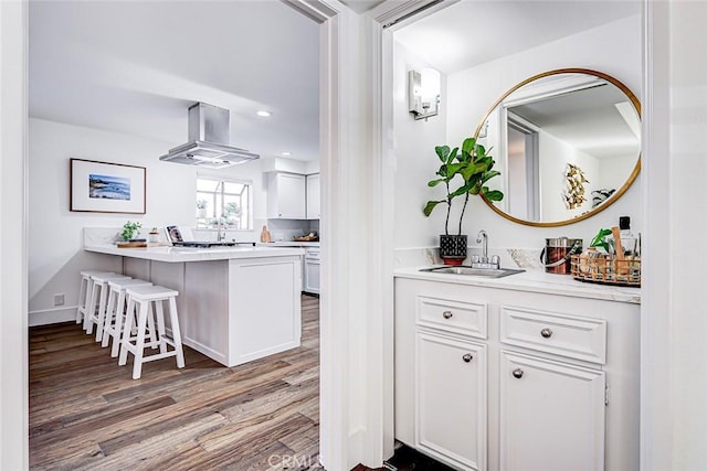 kitchen featuring a breakfast bar, a sink, wood finished floors, wall chimney exhaust hood, and white cabinets