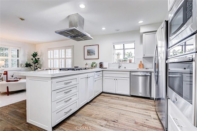 kitchen featuring light wood-type flooring, appliances with stainless steel finishes, a peninsula, island range hood, and light countertops