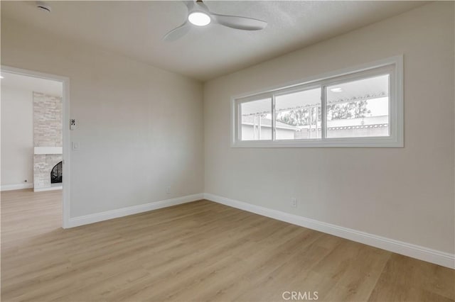 empty room with a stone fireplace, baseboards, light wood-type flooring, and ceiling fan