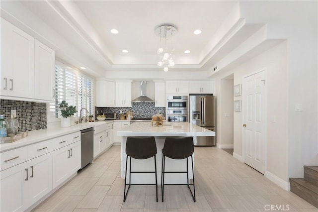 kitchen featuring wall chimney range hood, a breakfast bar, a tray ceiling, appliances with stainless steel finishes, and white cabinetry