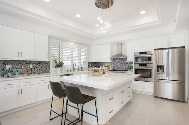 kitchen with a raised ceiling, wall chimney range hood, appliances with stainless steel finishes, and a center island