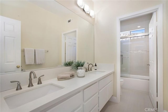 full bath with double vanity, toilet, bath / shower combo with glass door, and a sink