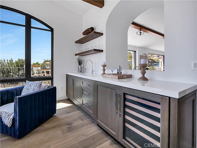 bar featuring beamed ceiling, light wood-style flooring, wine cooler, arched walkways, and indoor wet bar