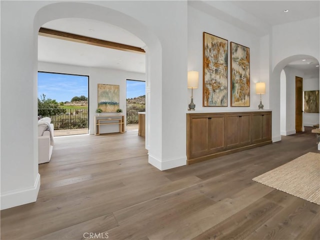 corridor featuring beam ceiling, wood finished floors, arched walkways, and baseboards