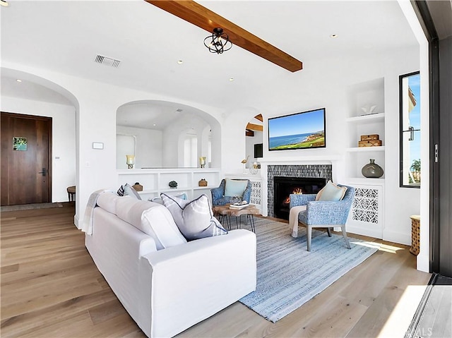 living room with built in features, visible vents, light wood finished floors, a tiled fireplace, and beamed ceiling