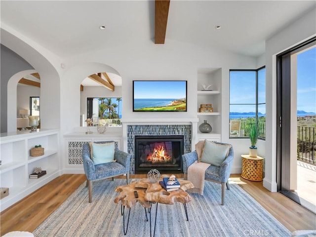 living area featuring built in features, lofted ceiling with beams, a tile fireplace, wood finished floors, and arched walkways