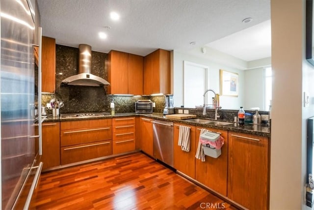 kitchen with a sink, stainless steel appliances, brown cabinetry, and wall chimney range hood