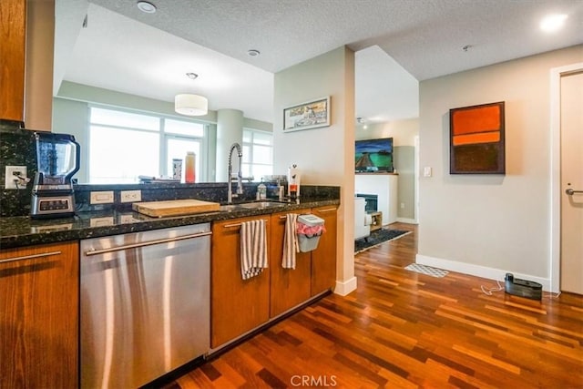 kitchen with brown cabinetry, dark wood finished floors, dark stone counters, a sink, and stainless steel dishwasher