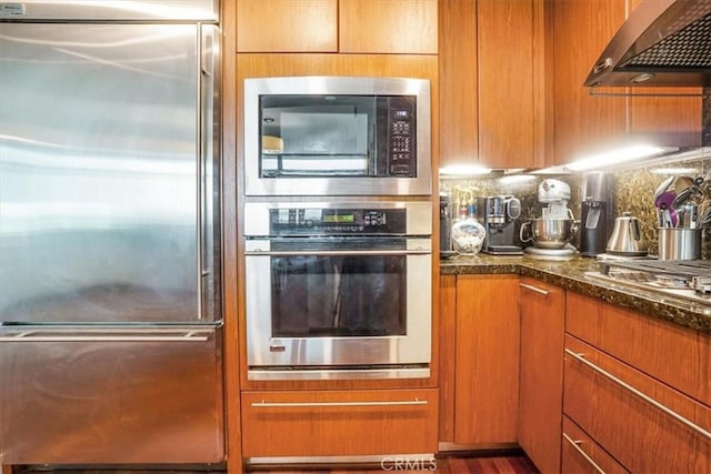 kitchen featuring range hood, dark stone countertops, tasteful backsplash, and built in appliances