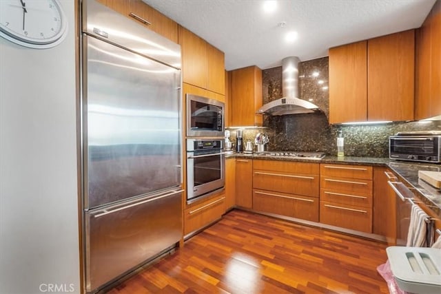 kitchen with dark wood-type flooring, a toaster, built in appliances, decorative backsplash, and wall chimney exhaust hood